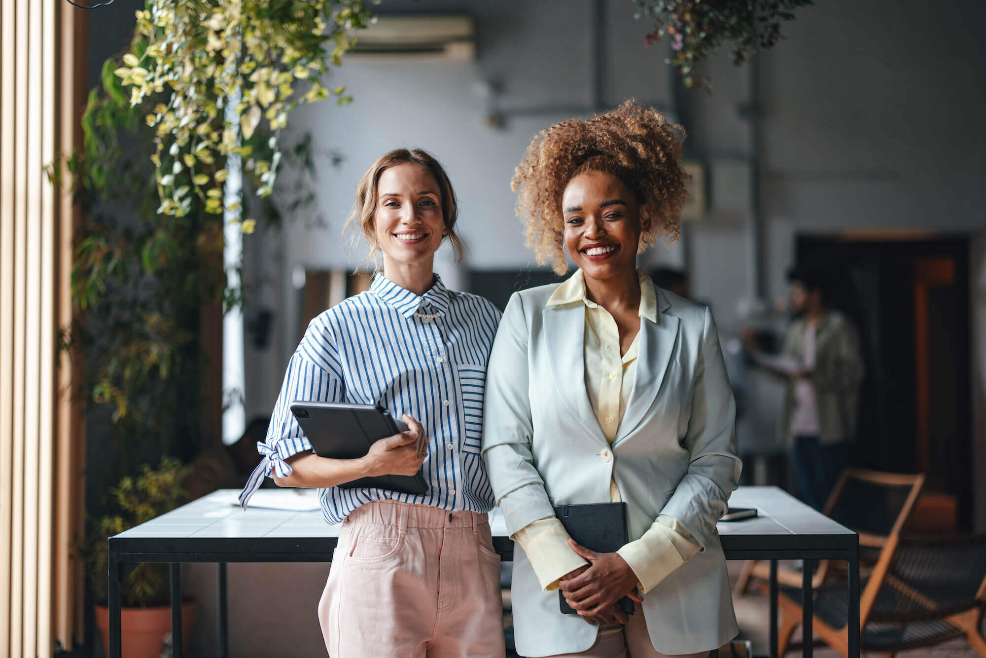 Two Happy Beautiful Businesswomen Looking At Camera While Standing In The Office